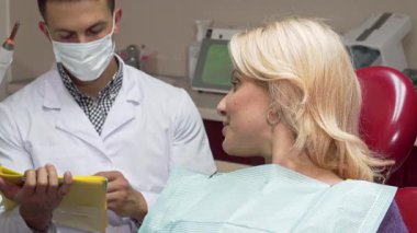 Neşeli, olgun bir kadın, parlak bir klinikte pozitif ışıldayan dişçi koltuğundan başparmağını doğruluyor. Bu arada, özenli erkek dişçisi tıbbi evrakları doldurup sıcak bir güven ve bakım ortamı yaratıyor.