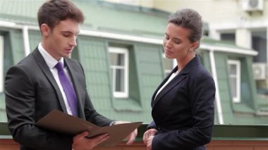 Sivri takım elbiseli genç bir adam, her ikisi de güneşli bir balkonda canlı bir iş görüşmesi yapan odaklanmış genç bir kadına fikirler sunuyor. Mesleki kıyafetleri ve özenli ifadeleri, modern çağda işbirliği ve yeniliğin enerjisini vurguluyor.