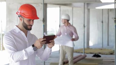 Turuncu bir şapka takan genç beyaz bir mimar, elinde dijital bir tablet tutarken, inşaat alanını incelerken güvenle gülümsüyor. Arka planda, bir mühendis arkadaşımız planlar üzerinde çalışıyor, ekip çalışmalarını ve çalışmalarını sergiliyor.