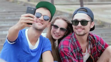 Üç genç arkadaş, bir erkek ve bir kadın, güneşli basamaklarda neşeyle selfie çekiyorlar, mutluluk ve dostluk saçıyorlar. Şık güneş gözlükleri ve günlük kıyafetleri kaygısız bir yaz havasını yansıtıyor. Bu anı arkadaşlığın değerli bir hatırası yapıyor.