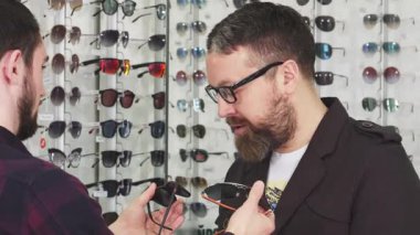 Olgun sakallı bir adam genç bir erkek optometristin parlak, modern bir gözlük dükkanında sunduğu iki gözlüğü incelikle inceliyor. Arka plandaki canlı gözlük görüntüsü alışveriş deneyimini güçlendiriyor.