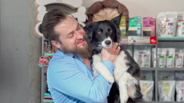 Şık sakallı neşeli bir adam neşeli köpeğini yanında tutuyor, canlı bir evcil hayvan dükkanında mutluluk saçıyor. Sevimli köpek, parlak kürkü ve sallanan kuyruğuyla, dostluk ve aşkın ılık atmosferine katılır.