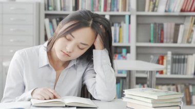 Yorgun Asyalı genç bir kadın kütüphanedeki çalışma masasında uyukluyor. Etrafı kitaplarla çevrili. Huzurlu atmosfer, sınavlara hazırlanırken uyanık kalma mücadelesini vurguluyor, akademik hayatın zorluklarını somutlaştırıyor