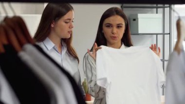 Canlı bir sahne, iki genç bayan moda mağazası çalışanının renkli kıyafetlerle çevrili şık bir beyaz tişörtü tartışmalarını canlandırıyor. Takım çalışmaları ve yaratıcılıkları, bu modaya uyum sağladıkları için parlıyor.