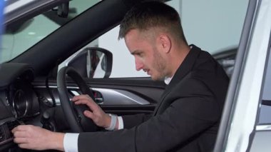 Gösterişli bir takım elbise giymiş odaklanmış bir işadamı, modern bir arabanın sürücü koltuğunda oturur ve yüksek teknolojili iç hatlarını dikkatle inceler. Bu büyüleyici sahne, yeni bir araç almanın heyecanını vurguluyor.