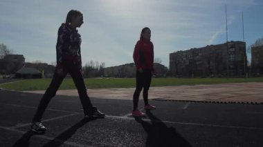 Canlı atletizm kıyafetleri giymiş iki genç kız güneşli pistte dinamik bacak ısıtma egzersizleri yapıyor ve kaslarını yaklaşan yarışa hazırlıyorlar. Sahne, odak ve kararlılıklarını yakalıyor, enerjilerini ve heyecanlarını gösteriyor.
