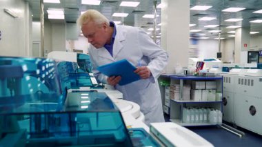 Beyaz laboratuvar önlüklü, beyaz saçlı seçkin bir adam elinde bir pano bulunan gelişmiş laboratuvar ekipmanlarını özenle inceliyor. Odaklanmış ifadesi modern bir bilimsel tesisin hareketli ortamında hassasiyete olan bağlılığını yansıtıyor.