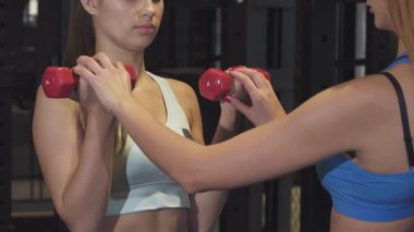 Odaklanmış bir kadın fitness koçu, kadın müşterisine canlı kırmızı dambıllar kaldırırken, destekleyici ve profesyonel bir atmosfer sergileyerek elle rehberlik eder. Bu güçlendirici an, takım çalışmasının ve motivasyonun önemini vurgular.
