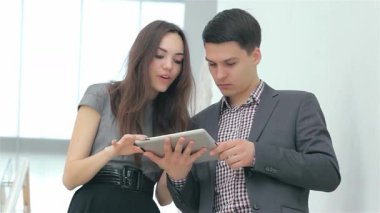 Memnun genç bir çift, şık takım elbiseli bir adam ve şık giyimli bir kadın, iş sonuçlarını hevesle bir tablette gözden geçiriyorlar. İfadeleri heyecan ve takım çalışması yayıyor, modern ofis ortamında başarının canlı enerjisini gösteriyorlar.