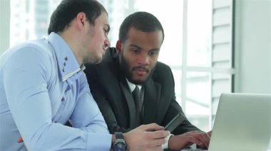 Biri açık mavi gömlekli, diğeri keskin siyah takım elbiseli iki kendine güvenen işadamı, bir dizüstü bilgisayar üzerinde işbirliği yaparak parlak ve modern bir ofiste fikir ve görüşlerini paylaşıyorlar. Gülüşleri takım çalışmasının ve başarının pozitif atmosferini yansıtıyor.