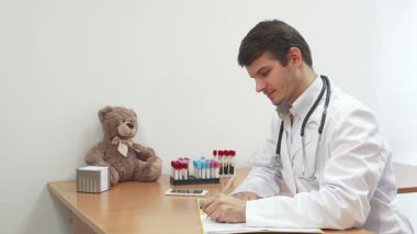 Parlak, hoş geldin ofisinde koyu saçlı ve sakallı bir erkek doktor not defterine notlar yazarken kameraya gülümsüyor. Çekici bir oyuncak ayı yanında oturuyor ve renkli test tüplerinin ima ettiği gibi sahneye biraz sıcaklık katıyor.