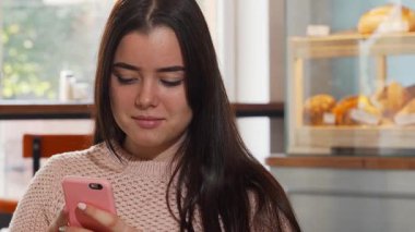 Uzun, dalgalı saçları olan neşeli genç bir kadın akıllı telefonuna bağlanmış, rahat bir kahve dükkanında mutluluk saçıyor. Arka planda lezzetli hamur işleriyle vurgulanan ılık ortam, sabah keyfi için mükemmel bir ortam yaratıyor.