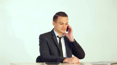 Sivri bir takım elbise giymiş genç bir işadamı, bir elinde kalem tutarken gülümsüyor ve cep telefonuyla konuşuyor, özgüven ve profesyonellik yayıyor. Masanın üzerindeki dizüstü bilgisayar ve tabletle temiz ofis ortamı,