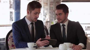 Şık bir kafede, iki resmi giyinmiş adam kalabalık bir restoranın sıcak atmosferiyle çevrili bir tableti incelerken derin bir tartışmaya daldılar. Özgüvenli ifadeleri ve profesyonel kıyafetleri başarılı bir ortaklığı yansıtıyor.