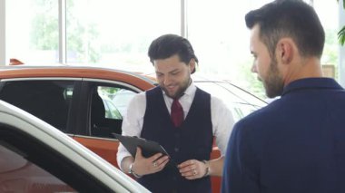 Şık giyinmiş karizmatik bir erkek araba satıcısı, parlak bir galeride potansiyel bir alıcıyla hevesle seçenekleri tartışıyor. Şık otomobillerle dolu modern sergi salonu, yeni araçları keşfetmek için davetkar bir ortam yaratıyor.