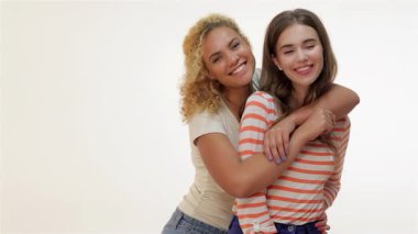 Biri kıvırcık sarı saçlı, diğeri uzun kahverengi saçlı iki neşeli kız arkadaş, yumuşak beyaz bir arka plana karşı sıcak bir kucaklaşmayı paylaşıyorlar. Işıl ışıl gülüşleri ve neşeli enerjileri arkadaşlığın özünü ve gençlik neşesini yansıtıyor.