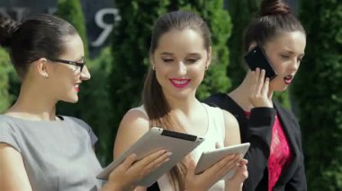 Üç kendine güvenen iş kadını açık havada duruyor, her biri kendi sohbetlerine dalmış, profesyonellik ve yoldaşlığın canlı bir karışımını sergiliyor. Şık kıyafetleri ve çekici ifadeleri modern bir şirket atmosferini yansıtıyor.