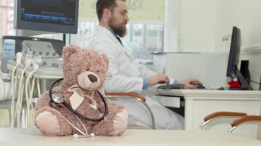 Şefkatli bir erkek çocuk doktoru MRI taramasını dikkatle incelerken steteskoplu peluş bir oyuncak ayı ön planda oturup çocuk sağlığı hizmetini sembolize ediyor. Bu iç açıcı sahne, pediatrik bakımın özünü yansıtıyor.
