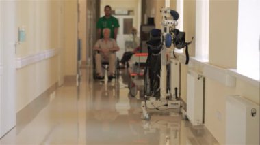Yeşil önlüklü kendini adamış genç bir doktor, tekerlekli sandalyedeki kıdemli beyaz bir adamı, parlak bir hastane koridorunda nazikçe itiyor. Sahne sıcaklık ve bakım saçıyor, tıbbi uzman ile hastası arasındaki bağı vurguluyor.