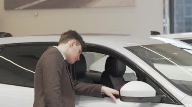 Olgun bir adam şık bir galeride sergilenen yeni bir arabanın içini incelemek için öne eğilir. Bu ilgi çekici sahne araba alışverişinin heyecanını yakalıyor, modern tasarımın cazibesini ve potansiyel sahipliğin heyecanını vurguluyor.