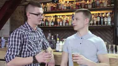 İki şık adam, canlı bir barda, neşe ve dostluk saçan soğuk bir bardak birayla kadeh kaldırıyor. Açık gri polo giyen sarışın adam ve kareli tişörtlü esmer adam bir dizi renkli şişelerle çevrili neşeli bir anı paylaşıyor.