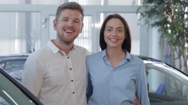 Neşeli esmer bir kadın, gülümseyen ortağıyla birlikte parlak bir galeride araba anahtarını gururla sergiliyor. Çift, yeni ve gösterişli araçlarla çevrili heyecan ve neşe saçıyor. Yeni başlangıçlarını ve ortak maceralarını simgeliyor.