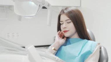 Uzun, koyu renk saçlı genç bir kadın diş ağrısından dolayı yanağına hafifçe dokunarak bir diş sandalyesinde oturuyor. Mavi bir diş önlüğü giymişti. Muayene olmayı bekliyordu. Diş kliniğindeki gerginlik ve endişeyle doluydu.