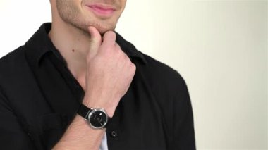 Çekici bir gülümsemesi olan yakışıklı genç bir adam, yumuşak beyaz bir arka plana karşı şık bir siyah gömleğe güvenerek çenesini tutuyor. Düşünceli ifadesi ve bakımlı görünüşü, parlak bir saatle tamamlanmış, davetkar bir davet yaratıyor.