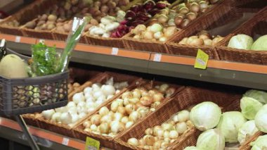 Neşeli genç bir çift hareketli hipermarkette taze ürünlerle dolu canlı bir tramvayı itiyor. Sahne, arka planda renkli sebzeler ve meyvelerle birlikte alışverişin heyecanını yakalıyor.