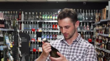 Çekici genç bir adam, hareketli bir süpermarket koridorunda şarap şişesini dikkatlice seçerken kameraya ışıldıyor. Onun kendine güvenen gülümsemesi ve şık ekose gömleği canlı bir alışveriş deneyimini yansıtıyor, şarap seçimini heyecan verici bir macera gibi hissettiriyor.