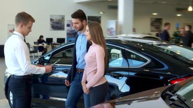 Modern bir bayilikte, genç bir çift, gösterişli siyah bir arabanın yanında dururken bir sözleşmeyi inceleyen dost canlısı bir satıcıyla karşılaşır. Atmosfer, potansiyel alımlarını tartışırken heyecan ve heyecanla dolu.