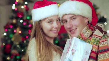 Sıcak süveterler ve şenlikli eşarplarla süslenmiş, sevgi dolu bir çift, sıcacık bir Noel odasında güzel paketlenmiş hediyeler tutarken neşeyle ışıldıyor. Yanıp sönen ışıklar ve canlı bir Noel ağacıyla çevrelenmiş şefkatli gülümsemeleri ışıldıyor.