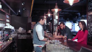 Canlı bir üçlü grup iki neşeli kız ve bir adam hareketli bir bar ortamında barmenden canlandırıcı bir şekilde içki sipariş ediyorlar. Kahkahalarla ve arkadaşlıkla kutlanan bar ışıklarının sıcacık parıltısıyla çevrili atmosfer heyecan vericidir.