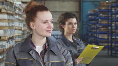 Koruyucu giysiler içinde ışıl ışıl genç bir kadın mühendis, pozitif ruhunu göstererek başparmak kaldırırken kendine güvenle ışıldıyor. Arka planda, meslektaşı titizlikle stokları kontrol ederek, canlı endüstriyel çalışmaların altını çiziyor.