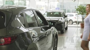 Uzun, dalgalı saçları olan çarpıcı bir kadın modern bir bayilikte parlak, şık bir araba tarafından büyülenir. Düşünceli düşüncelerini yansıtır. Parlak sergi salonu havası, onun potansiyel yeni aracının cazibesini arttırıyor.