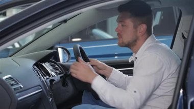 İyi bakımlı sakallı şık bir genç adam, lüks bir arabanın içini dikkatle inceliyor. Gösterge paneline odaklanmış, modern teknoloji ve tasarım özelliklerine olan ilgisini sergilerken,