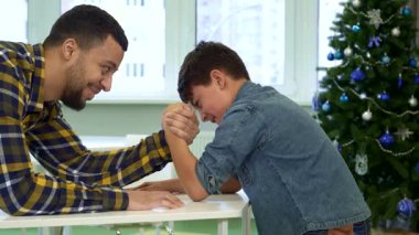 Neşeli bir baba ve küçük oğlu parlak bir masada neşeli bir bileklik güreşi yapıyorlar ve arka planda Noel ağacı parıldıyor. Çekici Afrikalı Amerikalı baba, kararlı oğlu zafer kazanırken gülümsüyor.