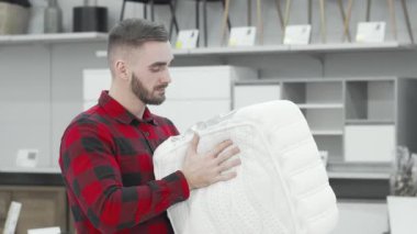 Kırmızı ekose gömlekli çekici bir sakallı adam modern bir mobilya mağazasında ortopedik yatak örneğini inceliyor. Gülümsemesi heyecan saçıyor. Konfor ve kalite dolu bir dünyada yol alırken bilinçli bir seçim yapıyor.