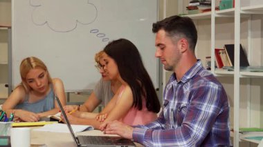 Yakışıklı genç bir erkek öğrenci, canlı bir çalışma grubu ortamında dizüstü bilgisayarı üzerinde çalışırken kendine güvenerek başparmağını kaldırıyor. Odaklanmış akranlarıyla çevrimiçi öğrenme, işbirliği ruhunu somutlaştırma, olumlu ve coşkulu bir şekilde yayılıyor.
