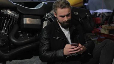 Şık sakallı neşeli bir beyaz adam yere oturmuş, akıllı telefonuna dalmış, güçlü bir helikoptere yaslanmış. Motosiklet atölyesi enerji ile uğulduyor, parlak krom detaylar ve aletler sergiliyor, motorcunun zevk aldığı gibi.