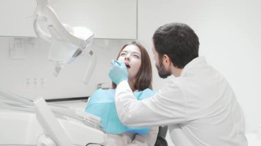 Genç bir kadın modern bir diş kliniğinde rahatça oturuyor, profesyonel bir dişçi olarak ağzı açık dişlerini titizlikle inceliyor. Parlak, steril ortam ve gelişmiş diş aletleri ağız temizliği ve bakımın önemini vurgular.