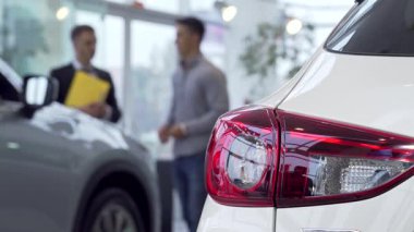 Canlı bir galeride, parlak araba ışıkları ön planda parlarken bir adam arka planda profesyonel bir satıcıyla seçenekleri tartışıyor. Bu dinamik sahne yeni bir araç satın almanın heyecanını yakalıyor.