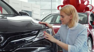 Kameraya sarı saç desenli neşeli genç bir kadın modern bir bayilikte yeni bir arabayı incelerken onaylıyor. Parlak aracın yanında dururken heyecanı parlıyor, yeni sahipliğin heyecanını ve...