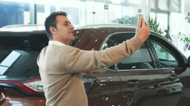 Parlak, parlak yeni bir arabanın önünde selfie çeken koyu renk saçlı şık bir adam. Bej bir kazak ve beyaz bir gömlek giyerek heyecan saçıyor, modern zarafet ve otomotiv cazibesinin mükemmel bir karışımını sergiliyor.