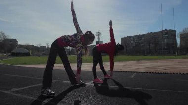 İki genç kız, yaklaşan yarışa hazırlanmak için güneş ışığında sırt ve omuz kaslarını ısıtarak dinamik esneme egzersizleri yapıyorlar. Canlı atletik kıyafetleri, açık mavi gökyüzüne karşı çok güzel tezat oluşturuyor.