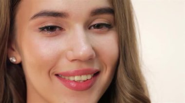 Parlak gülümsemesi ve kusursuz cildiyle genç beyaz bir kadının çarpıcı yakın plan portresi. Işıl ışıl gözleri ve yumuşak, dalgalı saçları yüzünü güzel çerçeveler, temiz beyaz bir arka plana neşeli ve pozitif bir titreşim yayar