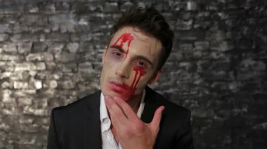 Saldırgan bir erkek vampir kendine güvenerek poz veriyor, eli nazikçe yüzüne dokunuyor, kan kırmızısı makyajla vurgulanıyor. Koyu tuğla duvar arka planı ürkütücü ama büyüleyici atmosferi güçlendiriyor. Cadılar Bayramı kutlamaları için mükemmel.