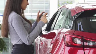 Uzun saçlı neşeli genç bir kadın akıllı telefonuyla meşgul olurken parlak bir şekilde gülümsüyor, modern bir galeride parlak kırmızı bir otomobilin yanında duruyor. Bu canlı sahne yeni bir araba seçmenin heyecanını yakalıyor, teknolojiyi harmanlıyor.