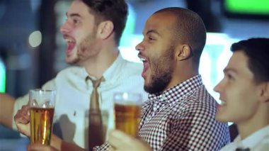 Üç hevesli arkadaş, çeşitli bir grup genç adam bardaklarını sevinçle kaldırarak barlarda heyecan verici bir futbol maçını kutluyorlar. Animasyon ifadeleri ve yoldaşlıkları heyecan verici bir atmosfer yaratıyor.