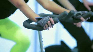 Hareketsiz bisikletler üzerinde güçlü bir şekilde pedal çeviren sporcuların yoğun odak noktalarını yakına çekerek, tonlu bacaklarını ve kararlılıklarını gösteriyor. Parlak spor salonu bu grup fitness seansının enerjisini arttırıyor.